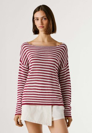 Jeune femme portant un pull rayé rouge et blanc à épaules dénudées et une jupe blanche asymétrique sur un fond uni.