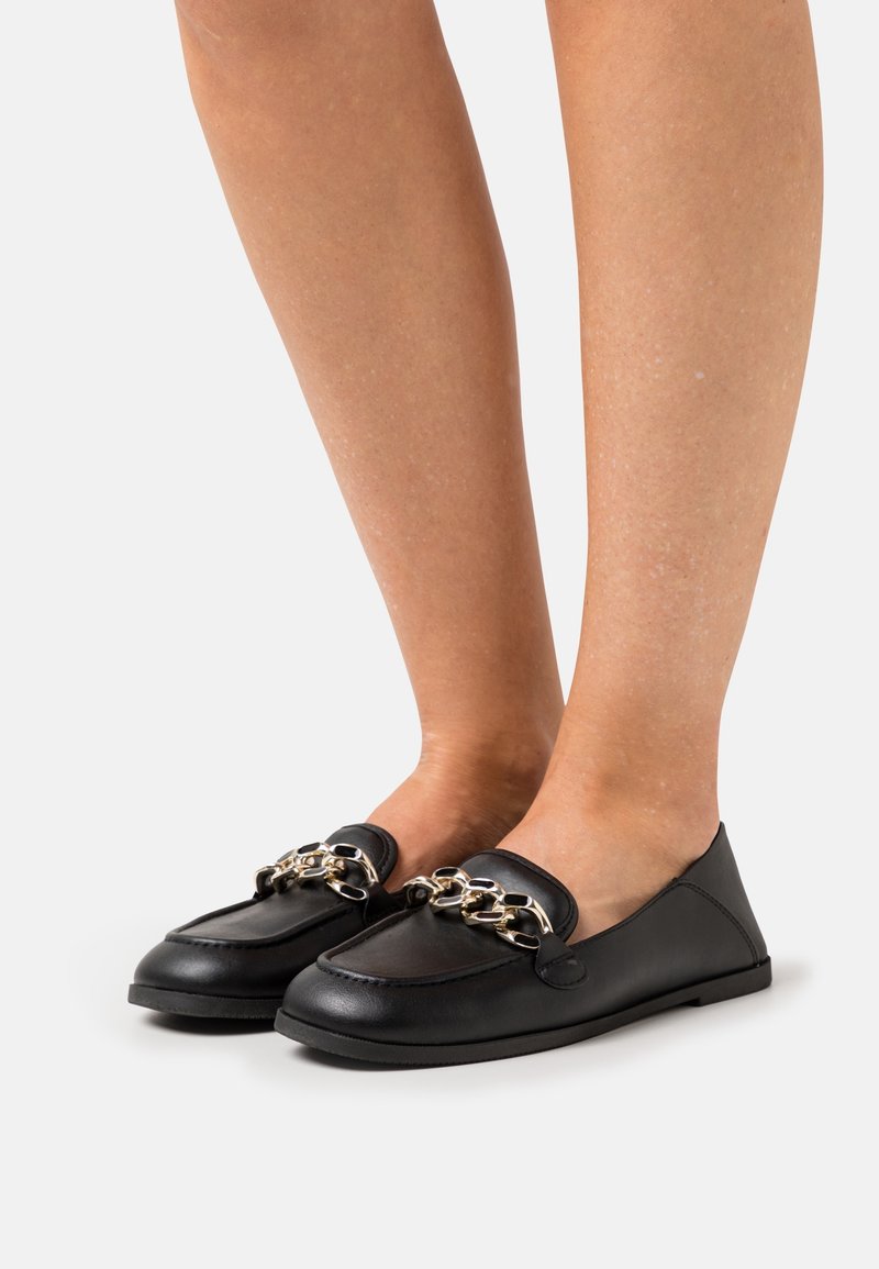 Jambes portant des mocassins en cuir noir avec un grand détail de chaîne en argent sur le dessus, debout sur un fond clair uni.