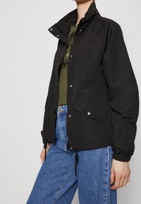 Kvinna klädd i en svart zip-up jacka över en olivgrön sweater och blå jeans, stående med en hand avslappnat vid sin sida.