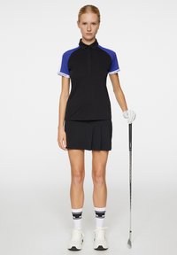 Kortärmad svart golfpolo med blå detaljer, tillsammans med en svart plisserad kjol. Vita sportstrumpor och golfklubba kompletterar outfiten.