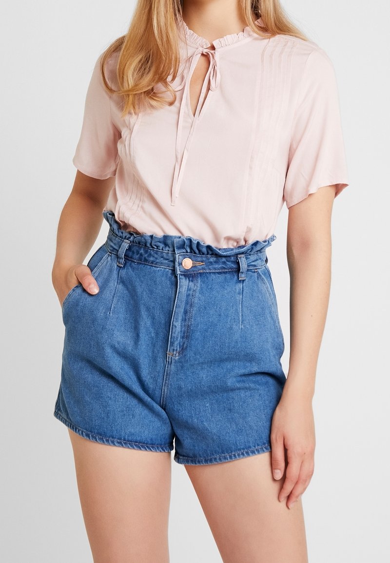 Blusa rosa claro con mangas cortas, escote con volantes y cierre con lazo, combinada con shorts de mezclilla azul de talle alto.