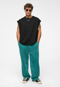 Zwarte oversized mouwloze t-shirt, teal zachte wijde broek, casual sneakers, gelaagde zilveren kettingen en zonnebril.