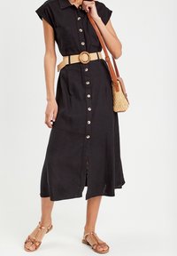 Femme portant une robe midi noire à boutons sur le devant avec une ceinture tressée beige, portant un sac bandoulière tressé marron clair, et des sandales plates marron.