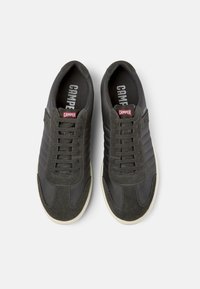 Un paio di sneaker nere Camper con accenti in suede, suole bianche, lacci e logo rosso Camper sulla linguetta, viste dall'alto su uno sfondo bianco.