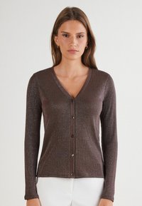 Eine Frau trägt eine taillierte, langärmelige, gebuttonte braune Glitzerstrickjacke mit V-Ausschnitt, kombiniert mit weißen Hosen, vor einem schlichten Hintergrund.