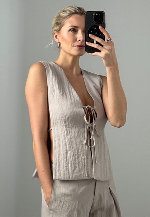 Kobieta z krótkimi blond włosami, ubrana w bezrękawnikowy beżowy lniany top z wiązaniami z przodu oraz dopasowane spodnie, robiąca selfie w lustrze za pomocą smartfona.