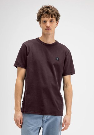 Jonge man met krullend haar die een effen donkerpaarse T-shirt en lichtblauwe spijkerbroek draagt, staand tegen een witte achtergrond.