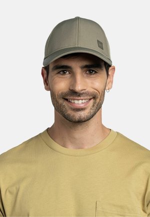 Mann mit kurzen dunklen Haaren, der eine hell olivgrüne Mütze, einen kleinen Creolen-Ohrring und ein senfgelbes T-Shirt mit Rundhalsausschnitt trägt und in die Kamera lächelt.