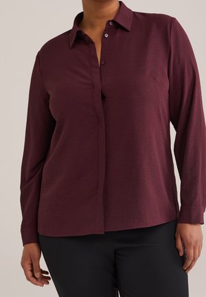 Kobieta ubrana w długą bluzkę w kolorze śliwkowym z guzikami i czarne spodnie, stojąca z ramionami swobodnie opuszczonymi wzdłuż ciała.