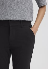 Čierne nohavice na mieru s hladkou textúrou, bočnými vreckami a stredne vysokým pásom, v páre so svetlosivým pleteným svetrom.