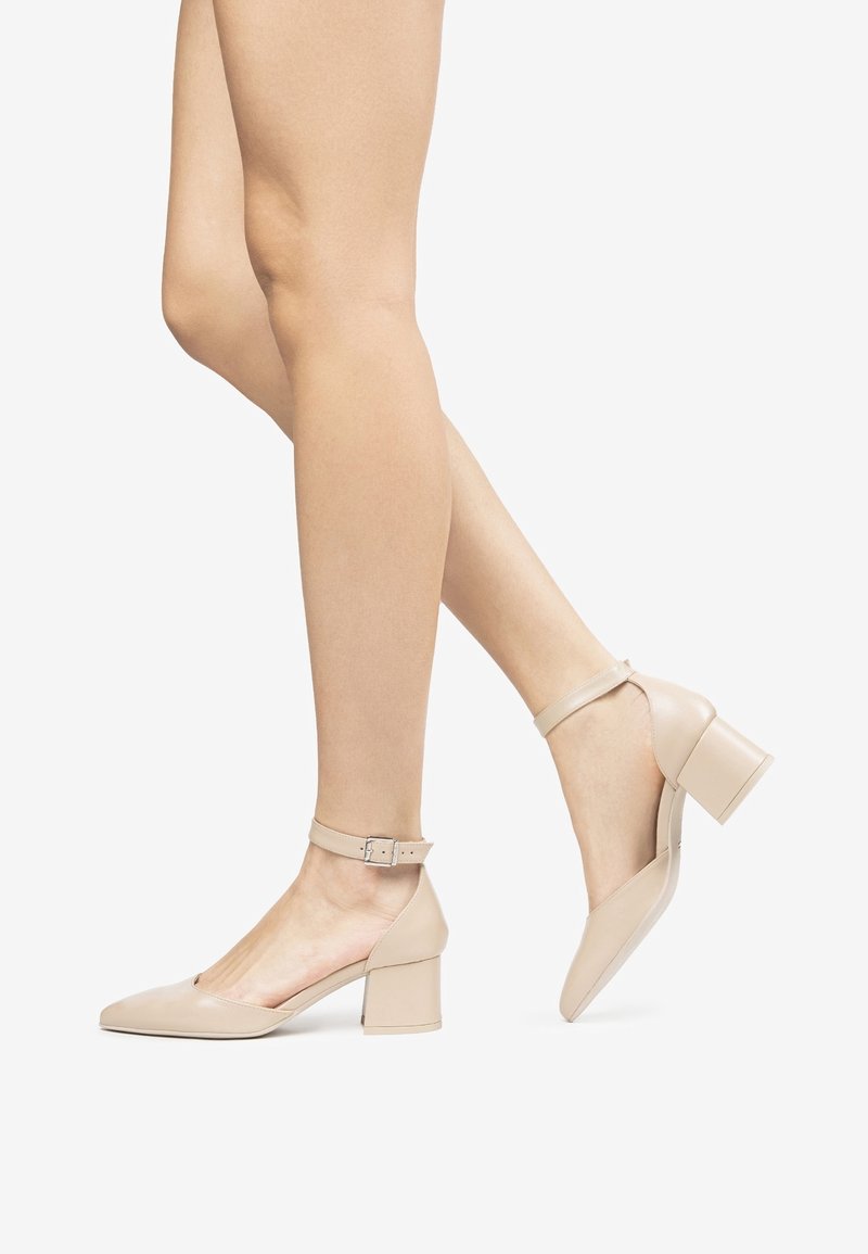 Jambes portant des chaussures beige à talon bloc fermé avec des brides à la cheville, un pied à plat et l'autre légèrement relevé sur fond blanc.