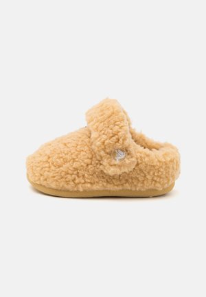 Blød beige fuzzy sutsko med en rem over toppen og et lille rundt logo på siden, sat mod en ensfarvet hvid baggrund.