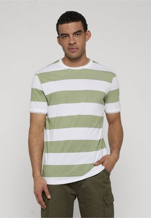 Mann trägt ein weißes und hellgrün horizontal gestreiftes Kurzarmshirt und olivfarbene Cargo-Hose, steht mit linker Hand in der Hosentasche.