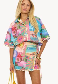 Tenue colorée comprenant une chemise à manches courtes et une jupe assortie. Fabriquée en tissu léger avec des imprimés graphiques tropicaux et des bords festonnés.