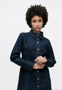 Φόρεμα από τζιν σε σκούρο μπλε με γιακά, μακριά μανίκια και μεταλλικά κουμπιά. Διαθέτει ορατές ραφές και εφαρμοστή σιλουέτα.