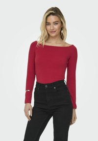 Crveni uski top dugih rukava s brojnim izrezom, uparen s crnim trapericama viskog struka. Značajan detalj: srebrna narukvica na zapešću.