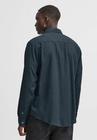 Camicia a maniche lunghe in tessuto verde scuro. Presenta un colletto, chiusure a bottoni ai polsini e un orlo leggermente curvato. Design semplice e texture.