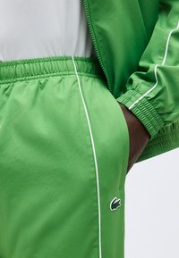 Κοντινό πλάνο ενός ατόμου που φοράει πράσινο παντελόνι και μπουφάν με λευκή ρίγα, με το χέρι στην τσέπη και το λογότυπο της μάρκας στο παντελόνι.