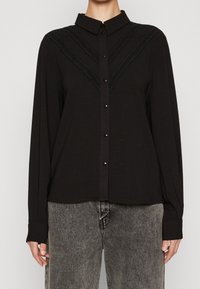 Black long-sleeve button-up shirt with a collar and textured accents in a V-pattern. Soft fabric, featuring glossy black buttons and a relaxed fit.