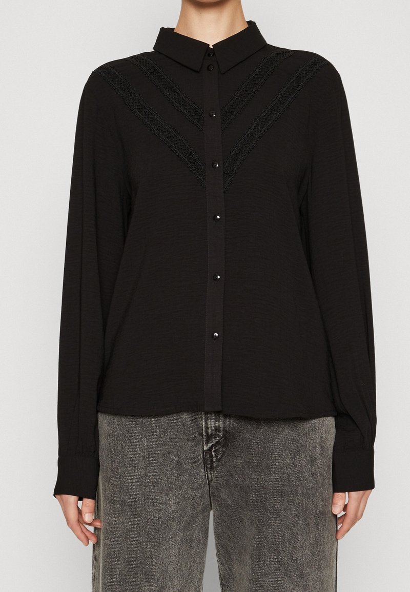 Black long-sleeve button-up shirt with a collar and textured accents in a V-pattern. Soft fabric, featuring glossy black buttons and a relaxed fit.