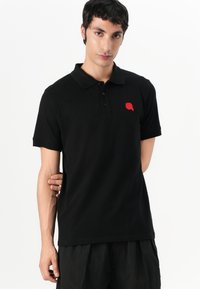 Μαύρο πόλο μπλουζάκι με κολάρο και κοντά μανίκια. Διαθέτει μια μικρή κόκκινη κεντημένη λεπτομέρεια στο στήθος. Απαλή ύφανση, κανονική εφαρμογή.