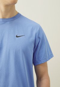 Camiseta de algodón azul con cuello redondo y mangas cortas, con un logo negro de Nike en la parte superior izquierda del pecho. Textura suave.