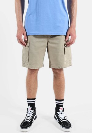 Homme portant un short cargo beige, des baskets montantes noires avec des lacets blancs, des chaussettes noires à rayures blanches et une chemise bleu clair.