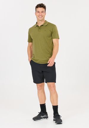 Lächelnder Mann trägt olivgrünes Poloshirt, schwarze Shorts, schwarze Socken und schwarze Sportschuhe, steht mit den Händen teilweise in den Taschen.