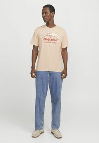 Lichtbeige katoenen T-shirt met een rode tekstontwerp met de tekst "JACK & JONES Surfer's Club", gecombineerd met blauwe rechte jeans en beige schoenen.