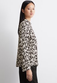 Schwarz-beige gemusterte Bluse mit Kragen, langen Ärmeln, entspannter Passform und Knopfmanschetten. Der Stoff wirkt glatt und leicht.