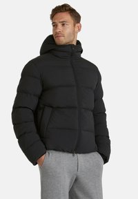 Giubbotto imbottito nero con zip frontale, cappuccio e tasche laterali. Caratterizzato da un design trapuntato e una superficie liscia. Abbinato a pantaloni sportivi grigi.