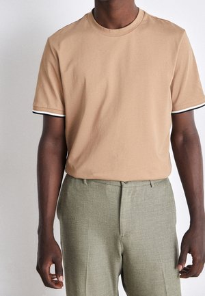 Hombre con camiseta de manga corta beige con ribete negro y blanco en los puños, combinada con pantalones grises claros de corte ajustado.