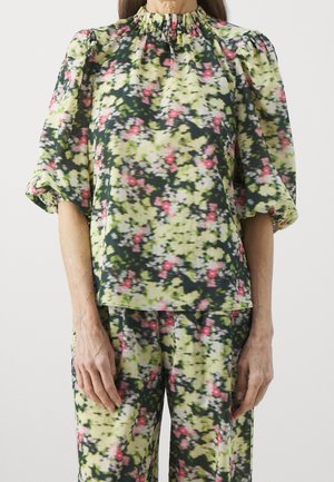 Frau trägt ein Oberteil mit Blumenmuster und Puffärmeln sowie passende Hosen in Grün, Gelb, Schwarz und Pink.