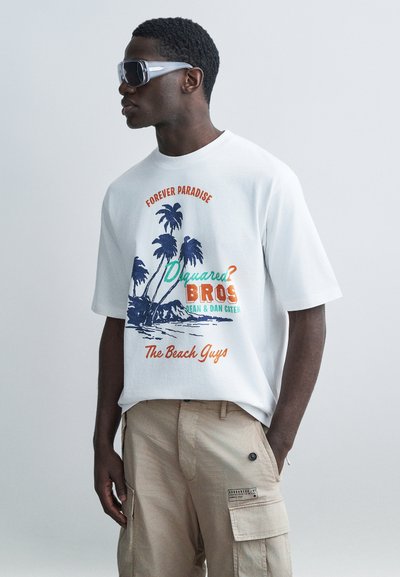 Νέος άνδρας που φοράει υπερμεγέθη λευκό t-shirt με γραφικό φοίνικα και κείμενο "Forever Paradise" και μπεζ παντελόνι cargo, ποζάροντας σε πλευρική όψη.