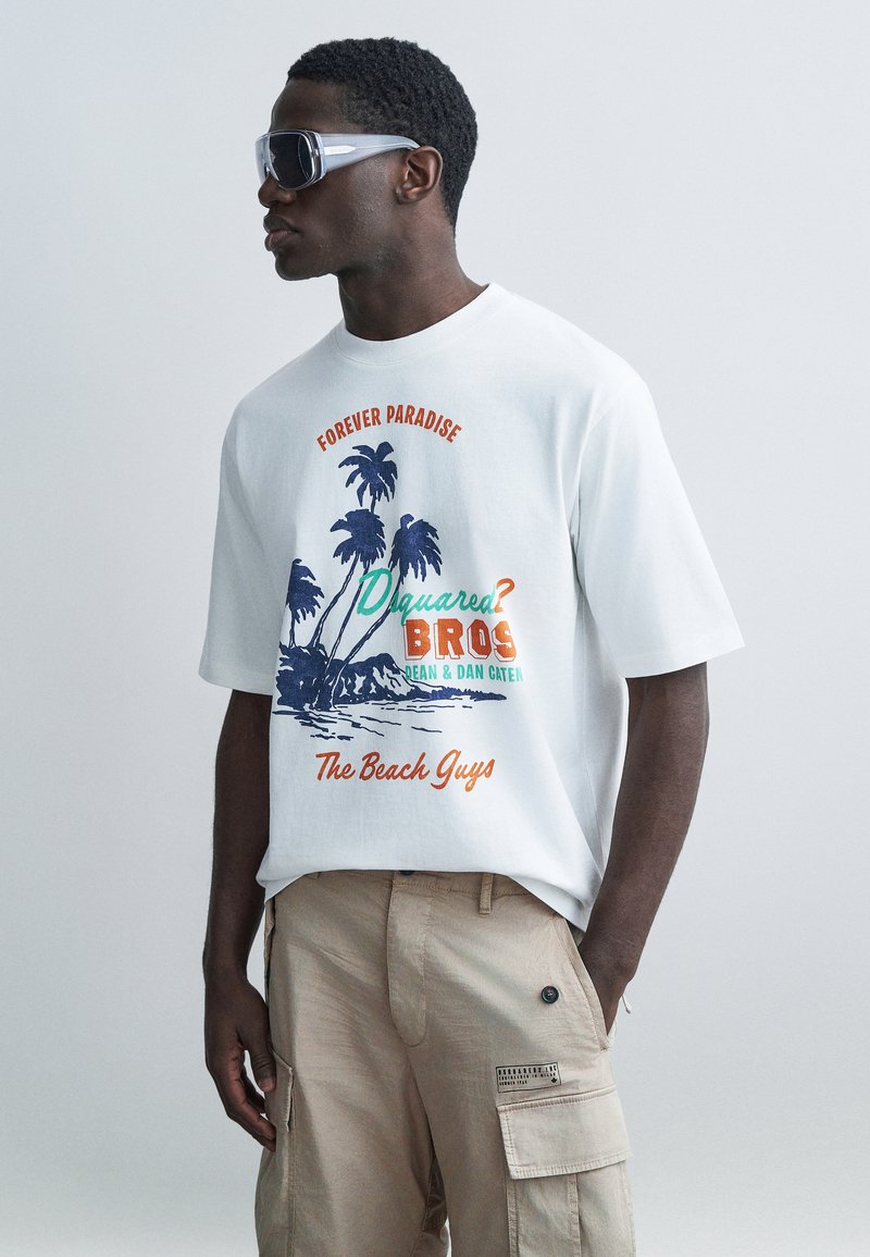 Junger Mann trägt ein übergroßes weißes T-Shirt mit Palmenmotiv und dem Schriftzug "Forever Paradise" sowie beige Cargo-Hosen, posiert in Seitenansicht.