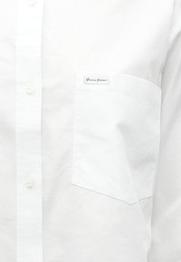 Valkoinen nappipaita, jossa vasemmassa rintataskussa "Guess Jeans" -merkki. Materiaali näyttää ryhdikkäältä ja sileältä.