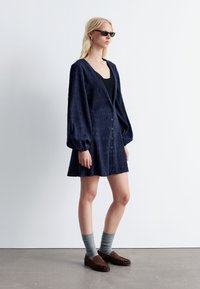 Dunkelblaues textiles Kleid mit V-Ausschnitt, langen Puffärmeln und Knöpfen an der Vorderseite. Ergänzt durch graue Socken und braune Slipper.