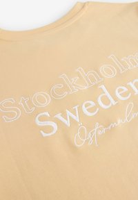 Žluté bavlněné tričko s bílým vyšitým nápisem: "Stockholm Švédsko Östermalm." Klasický kulatý výstřih, hladký materiál, ležérní design.