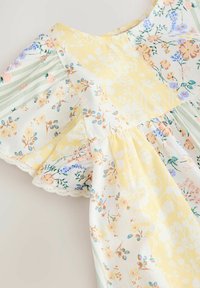 Bloemendess met gele en groene secties, met korte geraffineerde mouwen en een kanten rand aan de onderkant. Licht, zacht stoffen.