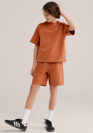 Kind in roestkleurig shirt met korte mouwen en bijpassende korte broek staat met één hand aan het hoofd, draagt witte sokken en zwarte sneakers.