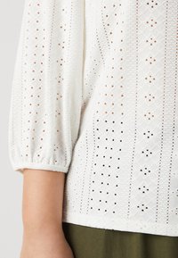 Balta blūze ar teksturētu acs audumu, ar puffētām piedurknēm un sarežģītu mazu izgriezumu rakstu. Zem blūzes ir redzama zaļa drēbe.