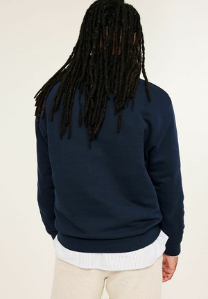 Persoon met lange dreadlocks, gekleed in een marineblauwe trui over een wit gestreept overhemd en beige broek, van achter gezien tegen een effen achtergrond.