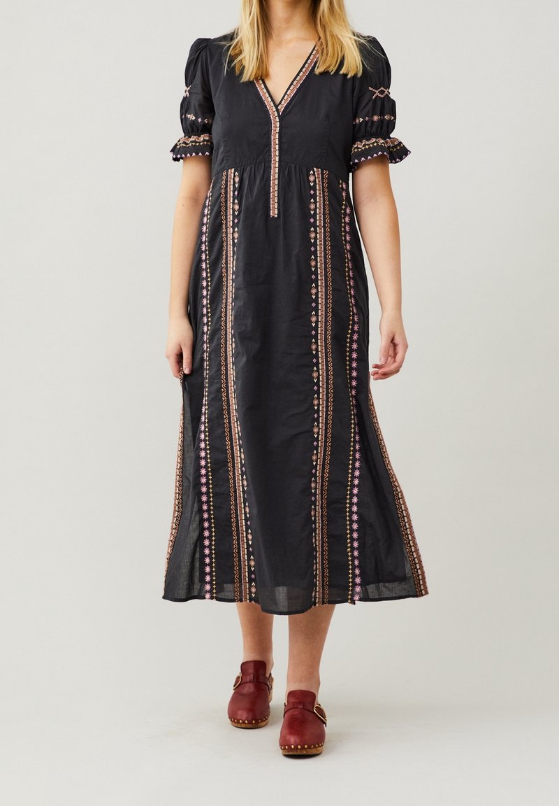Robe maxi noire à encolure en V, manches courtes bouffantes, avec une broderie colorée sur le devant et les côtés. Portée avec des sabots rouges.
