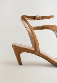 Sandal con tacco in suede marrone, con cinturino alla caviglia regolabile, punta aperta e tacco affusolato. Caratterizzato da una fodera interna liscia e una suola piatta.