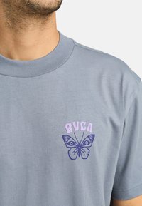 Camiseta de algodón gris con cuello redondo que presenta un gráfico de mariposa morada y el texto "RVCA" en morado claro encima del diseño.