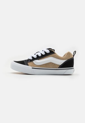Zapatilla de ante negra y marrón con cordones blancos, con una franja lateral blanca, acentos texturizados y una suela de goma con un acabado gris en la parte inferior.