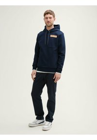 Sweatshirt bleu marine avec une poche frontale et une capuche à cordons, superposé sur une chemise blanche. Associé à un pantalon sombre et des baskets blanches.