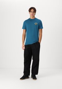 Teal cotton t-shirt with a round neck and a small gold logo on the chest, paired with black loose-fitting pants and black sneakers.