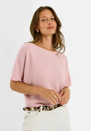 Femme portant un top en maille rose clair à manches courtes et un pantalon blanc avec une ceinture marron à motifs, posant les mains près de la taille sur un fond uni.