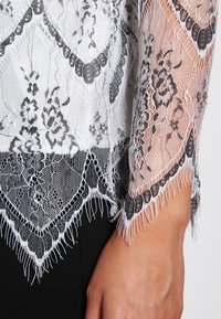Blusa in pizzo bianco e nero con motivi floreali, caratterizzata da bordi frangiati e maniche lunghe trasparenti, sovrapposta a una base nera solida.